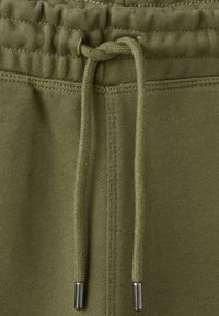 Primo piano della vita di un pantalone da tuta verde oliva con coulisse annodata e dettagli di cuciture visibili.