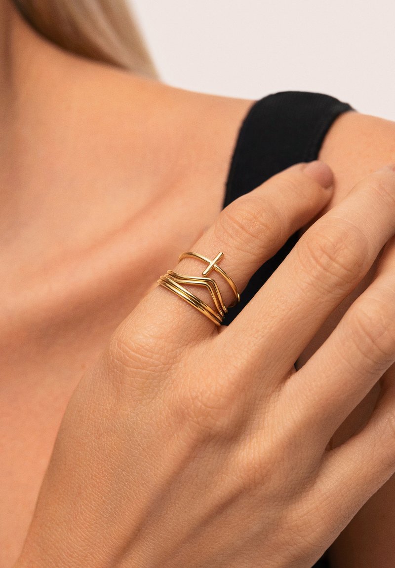 Gouden ringen hebben verschillende ontwerpen, waaronder een kruis, krommingen en gestapelde elementen, tentoongesteld op een hand tegen een neutrale achtergrond.