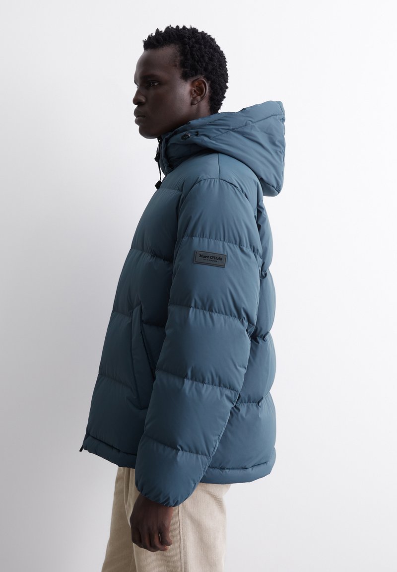 Türkisfarbene Pufferjacke mit Kapuze, die ein gestepptes Design und elastische Bündchen aufweist. Ein Logopatch ist am linken Ärmel sichtbar.