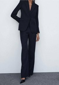 Blazer navy con un solo botón, solapas y dos bolsillos frontales, combinado con pantalones a juego. Unos zapatos de tacón altos negros completan el conjunto.