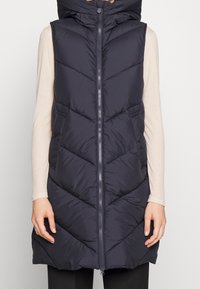 Chaleco acolchado azul marino con cremallera frontal y cuello alto; presenta costuras en forma de chevron y bolsillos laterales; diseño ligero e impermeabilizado.