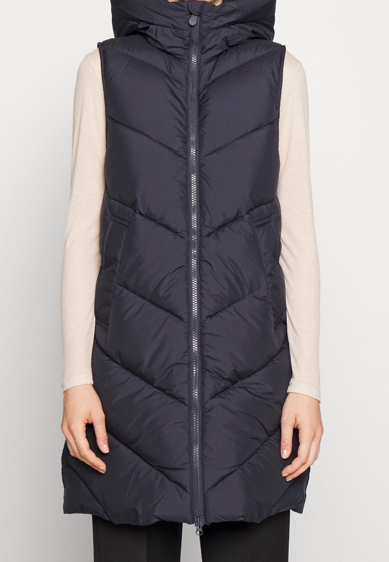 Chaleco acolchado azul marino con cremallera frontal y cuello alto; presenta costuras en forma de chevron y bolsillos laterales; diseño ligero e impermeabilizado.