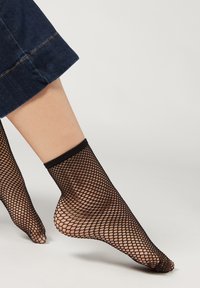 Sorte fiskenet ankelstrømper med et åbent vævemønster, der har en solid sort ribkant, båret med mørke denimbukser med opfoldede ben.