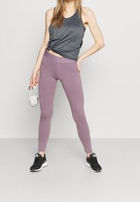 Graues sportliches Tanktop kombiniert mit helllavendelfarbenen Leggings. Die Leggings haben eine glatte Textur, eine enge Passform und einen breiten Bund. Schwarze Turnschuhe.