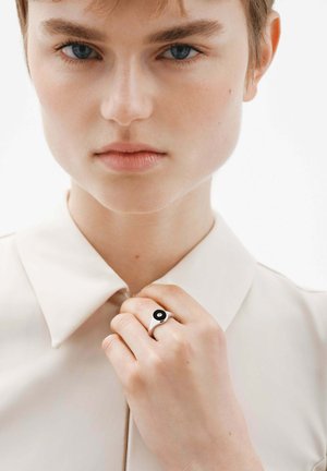 Anillo de plata con un acento circular negro, sobre una mano apoyada en una camisa color beige pálido con cuello. Piel suave y fondo neutro.