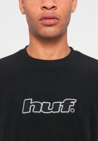 Sort t-shirt lavet af blødt stof, med et broderet hvidt "huf." logo i fed skrifttype hen over brystet. Standard rund halsudskæring.