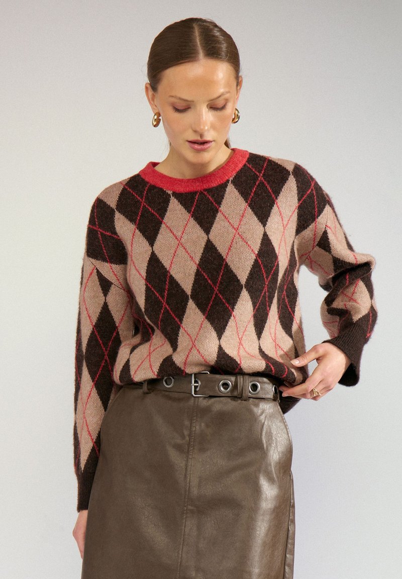Femme portant un pull à losanges marron et beige avec des touches rouges, associé à une jupe en cuir marron foncé et une ceinture noire avec œillets argentés.