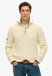 Crème kabelgebreide trui met een kwart-rits kraag, gedragen over een wit shirt. Blauwe spijkerbroek, bruine riem en een polshorloge maken de outfit compleet.