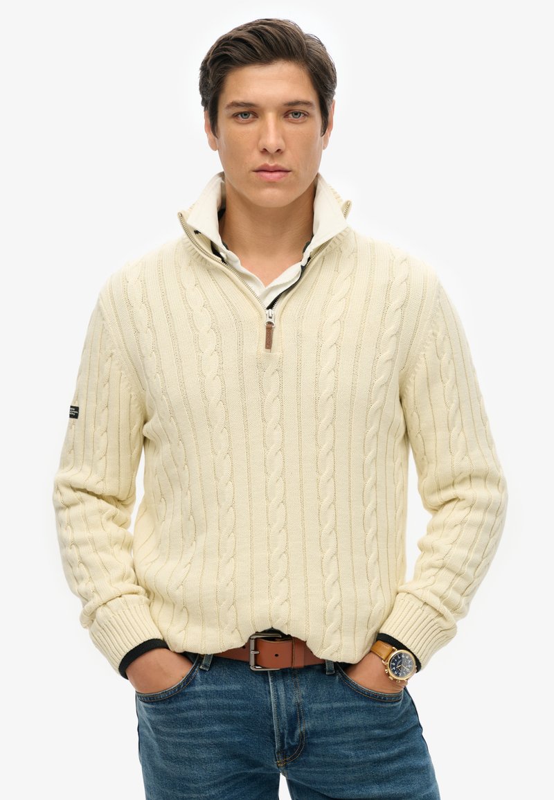 Crème kabelgebreide trui met een kwart-rits kraag, gedragen over een wit shirt. Blauwe spijkerbroek, bruine riem en een polshorloge maken de outfit compleet.