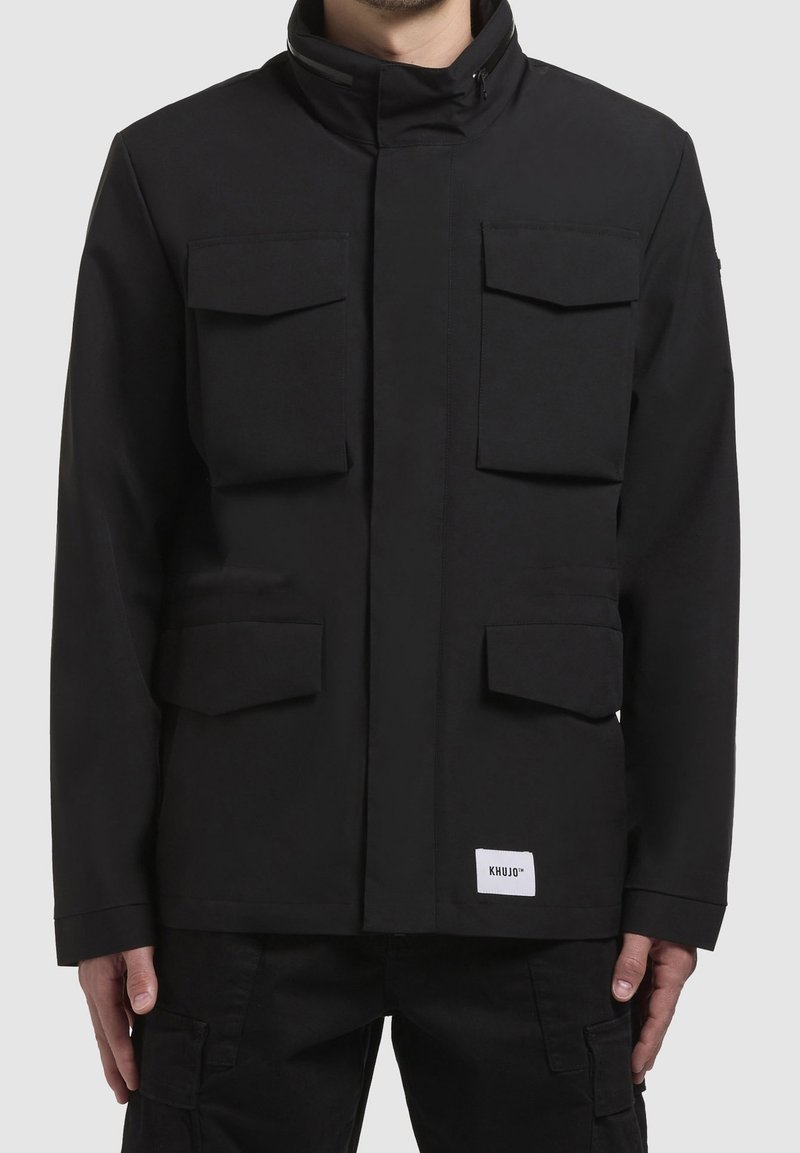 Homme portant une veste noire avec quatre poches avant à rabat et une petite étiquette blanche "KHUJO" en bas sur le devant.