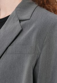 Γκρι σακάκι από υφασμάτινη υφή, με γιακά με εγκοπή και μία τσέπη στο στήθος, με εφαρμοστή γραμμή και λεία υφή.