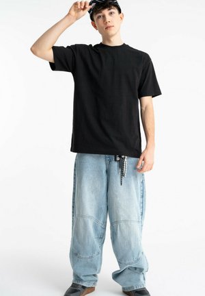 Młody mężczyzna poprawiający czarną czapkę, ubrany w luźną czarną koszulkę, obszerne jasnoniebieskie dżinsy z breloczkiem i czarne buty na tle białego tła.