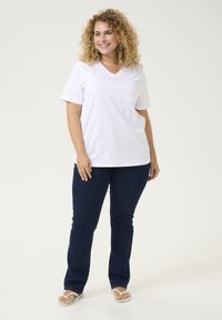 Camiseta de algodón blanca con cuello en V y mangas cortas, combinada con jeans de denim azul oscuro. El modelo lleva chanclas blancas. Un atuendo simple y casual.