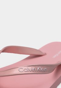 Ροζ σανδάλι τύπου σαγιονάρας Calvin Klein με διαφανείς λουριά που φέρουν ανάγλυφο το όνομα του бренδου, σε απλό λευκό φόντο.