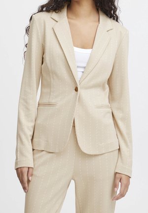 Femme portant un blazer beige à fines rayures avec un bouton et un pantalon assorti par-dessus un haut blanc, montrée du cou jusqu'au milieu de la cuisse.