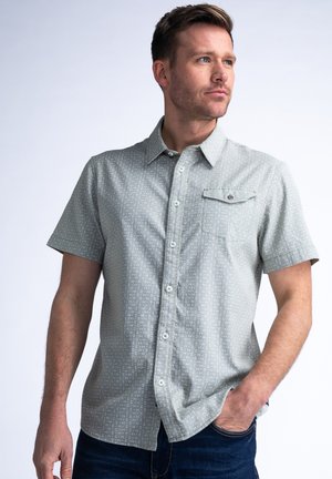 Camisa de manga corta gris claro con un sutil patrón geométrico, botón en la parte delantera y un bolsillo en el pecho con cierre de botón. Tela de mezcla de algodón.