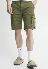 Olivgrüne Cargo-Shorts mit zwei Seitentaschen, aus Baumwollstoff, reguläre Passform und sichtbaren Nähdetails. Getragen mit weißen Socken und beigen Schuhen.