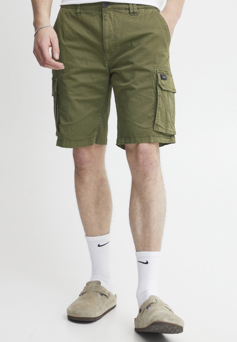 Olivgrüne Cargo-Shorts mit zwei Seitentaschen, aus Baumwollstoff, reguläre Passform und sichtbaren Nähdetails. Getragen mit weißen Socken und beigen Schuhen.