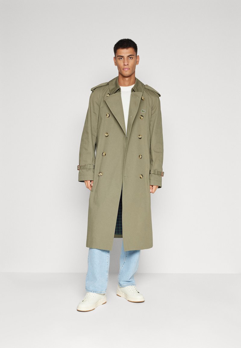 Olivgrüner Trenchcoat mit doppelter Knopfleiste, großen Revers, Schulterklappen und Knöpfen. Kombiniert mit hellblauen Jeans und weißen Sneakers.
