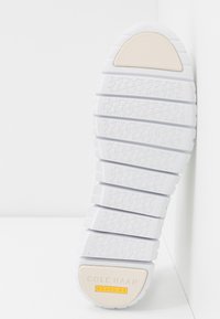 Semelle de chaussure en caoutchouc segmentée blanche avec grip texturé, coussinets beige au talon et aux orteils, et logo "Cole Haan Grand.OS" sur le talon.