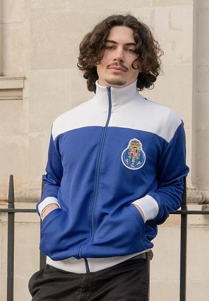 Blauwe en witte zip-up jacket met een hoge kraag en een logo aan de voorkant. Zacht materiaal met contrasterende witte mouwen en manchetten.