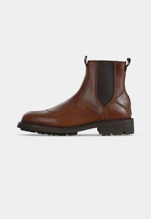 Botte Chelsea en cuir marron avec des empiècements élastiques noirs sur les côtés et une semelle crantée épaisse en caoutchouc noir sur un fond gris clair.