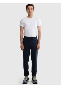 Vyras stovintis dėvintis baltą trumpomis rankovėmis marškinėlį, tamsiai mėlynas sportines kelnes su raišteliu ir mėlynas bei pilkas sportinius batelius ant vientiso fono.