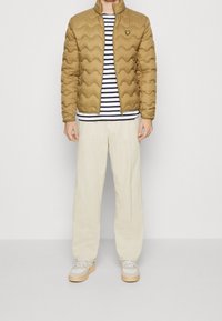 Hombre vistiendo una chaqueta acolchada color beis sobre una camiseta de rayas en blanco y negro, pantalones de pana color crema y zapatillas blancas con suelas de goma.