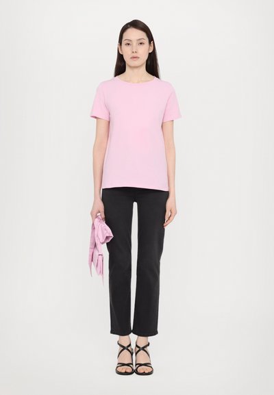 T-shirt rosa a maniche corte con scollatura rotonda, abbinato a pantaloni neri a gamba dritta. Il soggetto tiene una sciarpa rosa chiaro. Sandali neri con scarpette.