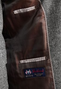 Intérieur de veste de costume en laine de Shetland avec une doublure en satin marron, deux petites poches, et une étiquette bleue avec un texte rouge indiquant la marque et l'origine.