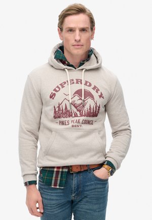 Beige hoodie met een grafische afbeelding van bergen en bomen. Gelaagd met een groen geruit overhemd, spijkerbroek en een bruine riem.