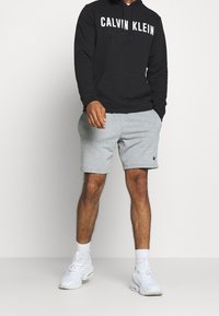 Hombre con sudadera negra de Calvin Klein, pantalones cortos grises claros de Nike, calcetines blancos y zapatillas deportivas blancas, de pie contra un fondo liso.