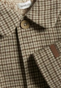 Brun ternet jakke lavet af blødt stof med en fyldig beige foring. Den har krave, knaplukning og læder mærke.