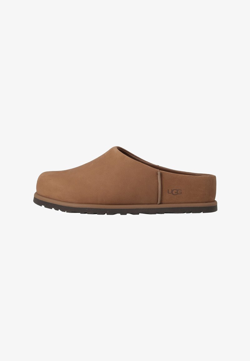 Καφέ παντόφλα UGG χωρίς κορδόνια με κλειστή στρογγυλεμένη μύτη και επίπεδη μαύρη σόλα, ορατό λογότυπο στο πλάι.