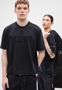 Tricouri negre din bumbac cu logo-ul „TOMMY JEANS” în relief, guler rotund și mâneci scurte; model purtând blugi închisi la culoare și având un rucsac negru.