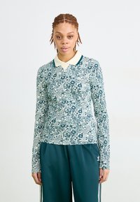 Žena s pletenom kosom nosi polo majicu dugih rukava s cvjetnim uzorkom i tamnozelenim Adidas hlačama za trčanje, stojeći protiv jednostavne pozadine.