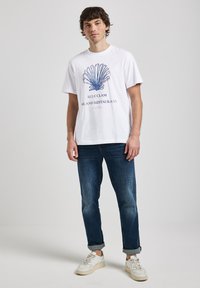 Bílá tričko s modrou grafikou mušli a nápisem "BLUE CLAM BAR AND RESTAURANT", spojené s tmavě modrými džíny a bílými teniskami.