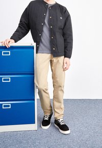 Zwart gewatteerd jack met gouden drukknoopjes, grijs T-shirt, beige rechte broek en zwarte suède sneakers. Blauwe metalen archiefkast op de achtergrond.