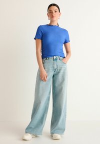 Modrá textúrovaná blúzka s krátkym rukávom a volánikmi, kombinovaná s nohavicami s vysokým pásom v svetlom umývaní a bielymi teniskami.