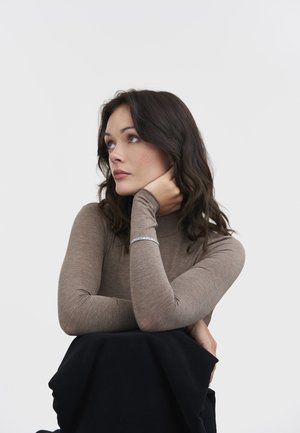 Woman with dark wavy hair wearing a brown long-sleeve top, resting chin on hand, and looking thoughtfully to the side against a plain background.