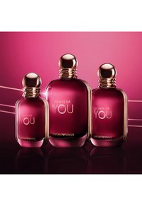 Tre bottiglie di profumo in vetro color bordeaux con tappi dorati e testi "Power of You" e "Emporio Armani" su sfondo rosa.