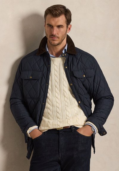 Uomo che indossa una giacca trapuntata blu navy sopra un maglione crema a trecce e una camicia azzurra con colletto, con le mani nelle tasche di jeans scuri.