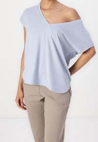 Ljusblå kortärmad topp med V-ringning och exponerade axlar, tillverkad av mjukt material. Löslig passform med rundad fåll, bärs tillsammans med beige byxor.