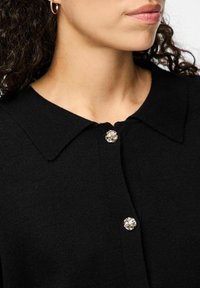 Črni kardigan z širokim ovratnikom, teksturirano tkanino in srebrnim dekorativnim gumbom na izrezu. Povečan posnetek.