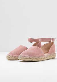 Różowe zamszowe espadryle z paskiem na kostkę, płaskimi plecionymi podeszwami z jutowego włókna i otwartymi bokami dla zapewnienia wentylacji.