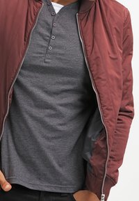 Mężczyzna w bordowej kurtce z suwakiem, rozpiętej nad ciemnoszarym, zapinanym na guziki podkoszulku typu henley, z rękami częściowo w kieszeniach czarnych spodni.