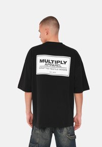 Mustat oversize T-paita, joka on valmistettu puuvillasta. Taakse on painettu suorakaiteen muotoinen valkoinen logo, jossa lukee "MULTIPLY APPAREL", ja paita on miellyttävän sileä.