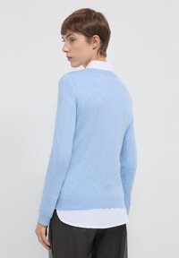 Maglione azzurro chiaro con maniche lunghe, scollatura a girocollo, texture morbida, indossato sopra una camicia bianca con colletto, con un taglio semplice e dritto.