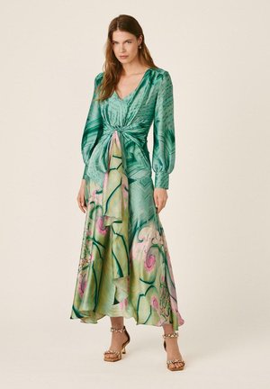 Mujer con un vestido cruzado de manga larga, verde y flores en tonos pastel, con mangas abullonadas y tacones con correa en el tobillo, de pie frente a un fondo liso.