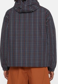 Person som bär en marinblå rutad jacka med huva och rostfärgade byxor, sedd bakifrån mot en enfärgad bakgrund.